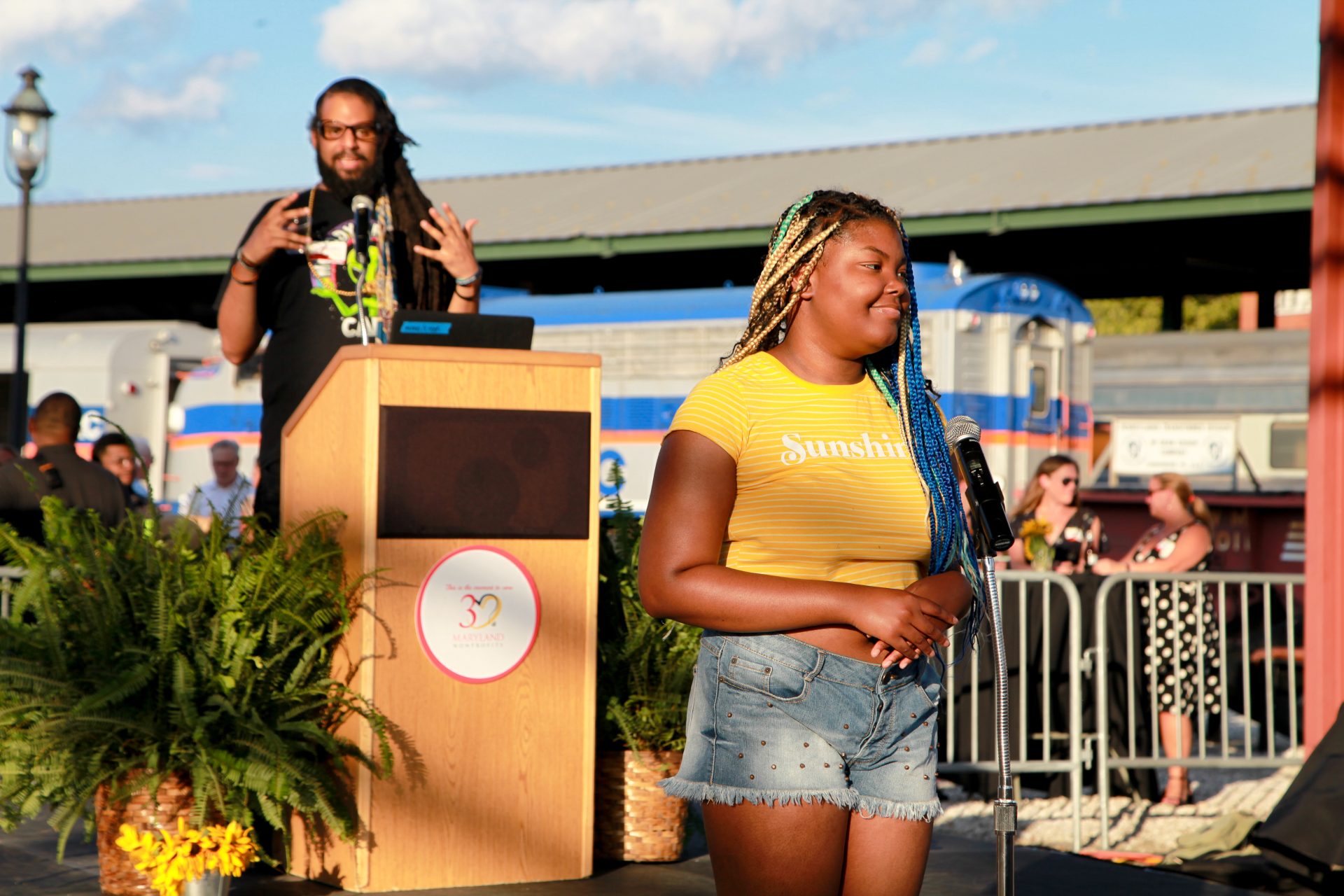 A young Black woman wearing a yellow shirt and denim shorts stands in front of a podum, smiling, ready to recite a poem
