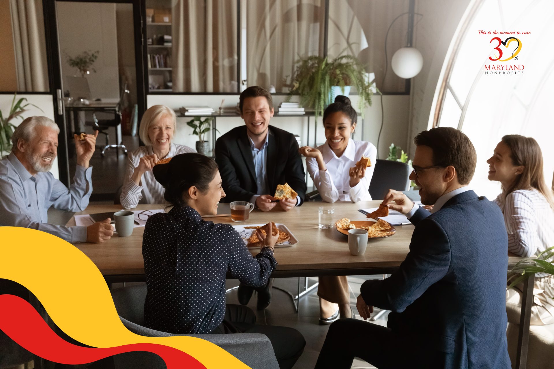 Group of people in work attire sit around a conference table eating pizza and laughing. They are diverse in age and race.